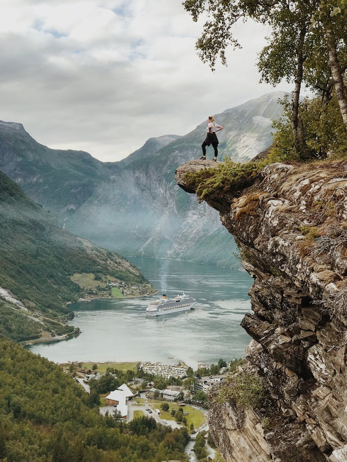services-02 Person enjoys breathtaking view from a cliff above Geiranger Fjord, Norway, with clear skies and cruise ship below.